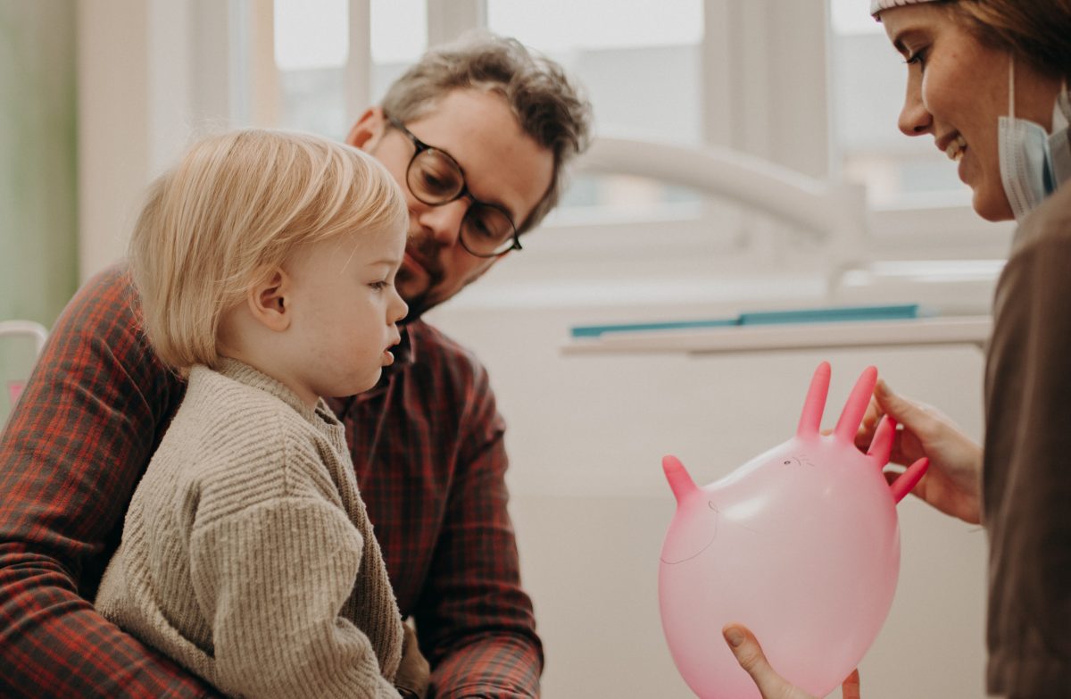 Kleinkind sitzt beim Zahnarzt und bekommt Luftballon auf Handschuh vom Zahnarzt in der Zahnarztpraxis Schnutenwerk in Kempen